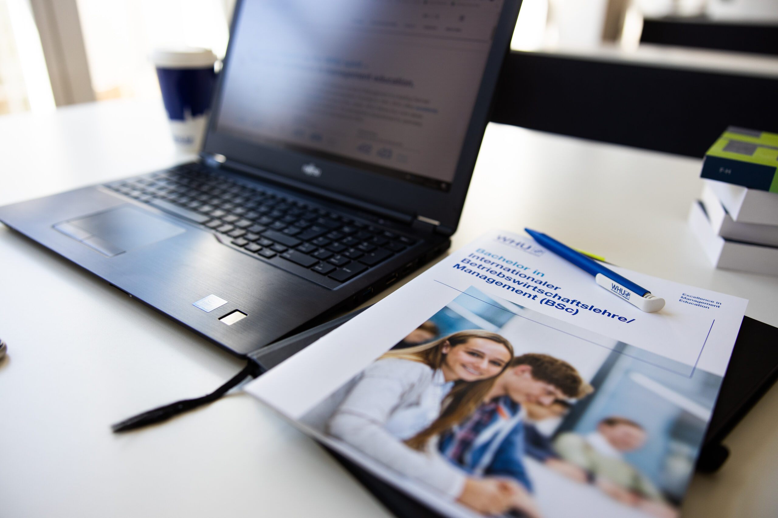 A laptop on a desk next to a WHU brochure and branded pen, with blurred books in the background.