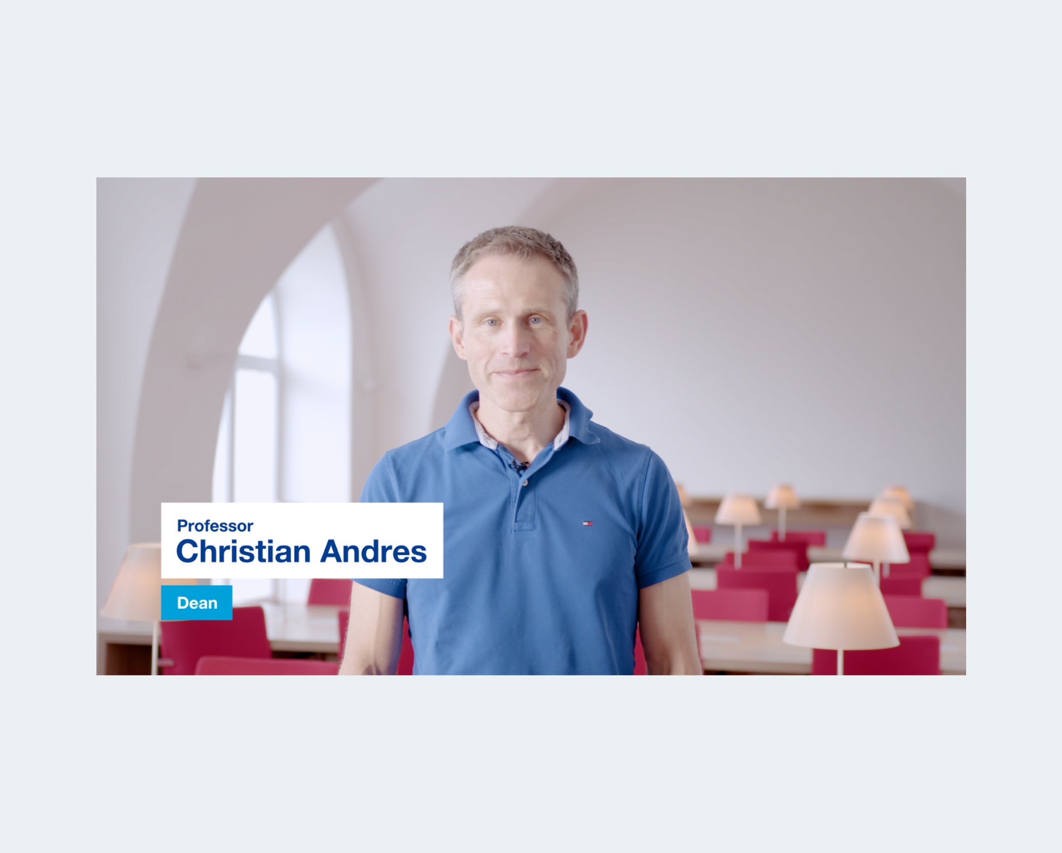 Man standing in a bright study space with red seating and table lamps, speaking in an interview-style video. A lower-third graphic displays his professional title.