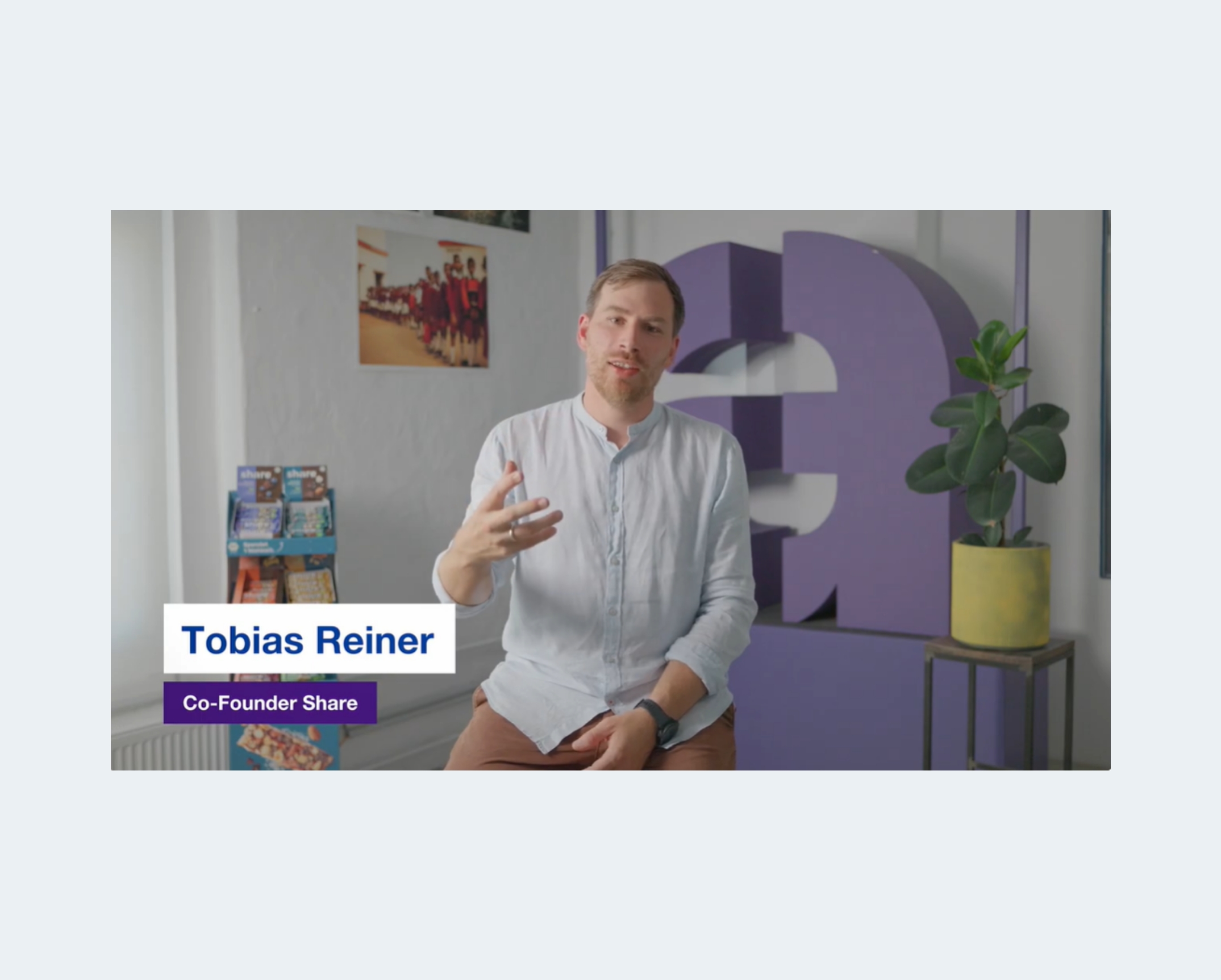 Interview-style video scene showing a person speaking while seated in front of a large decorative letter and product display.