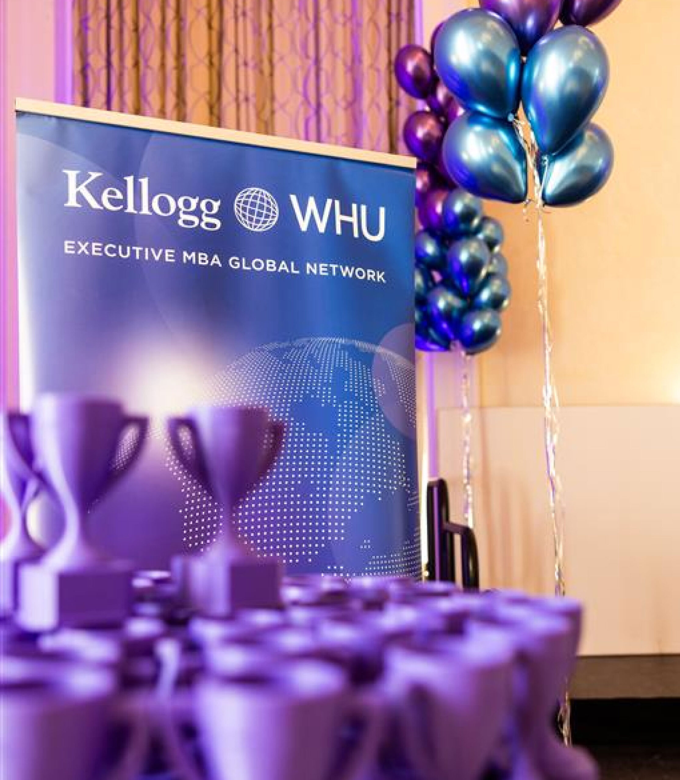 Kellogg-WHU Executive MBA event setup with branded banner, metallic balloons, and trophy awards in the foreground.
